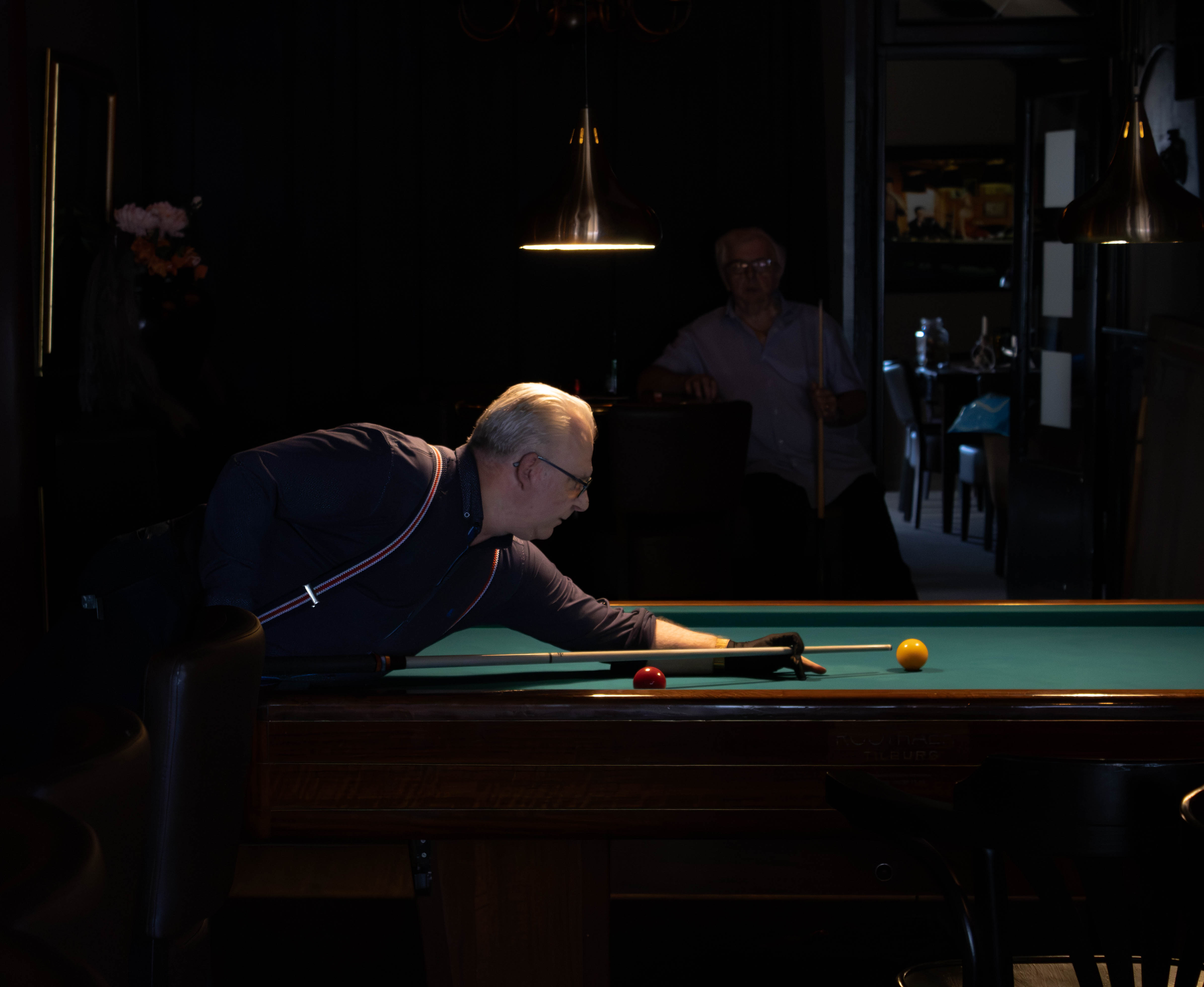 Een biljarter buigt over de tafel en richt zijn keu terwijl een andere man toekijkt